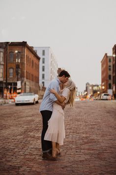 a man and woman kissing in the middle of a brick road with buildings behind them