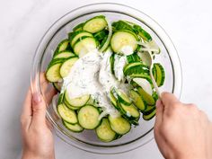 two hands holding a glass bowl filled with cucumber slices and sour cream sauce