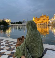 a woman sitting on the ground in front of a large body of water with a golden building behind her