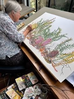 an older woman sitting in front of a painting on a table with paintbrushes