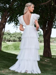 the back of a woman in a white wedding dress standing under trees with her hands on her hips