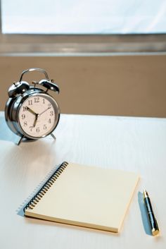 an alarm clock next to a notepad and pen on a white table with a window in the background