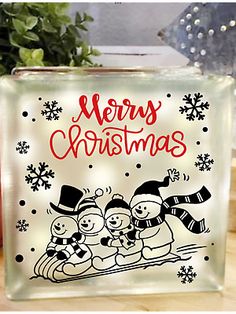 a glass block with a merry christmas scene on the front and bottom, sitting next to a potted plant