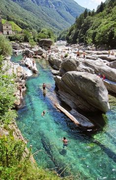 people are swimming in the water near some rocks and trees, while another person is standing on one side of the river