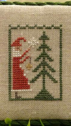 a cross - stitch christmas scene with a santa clause holding a pine tree in front of it