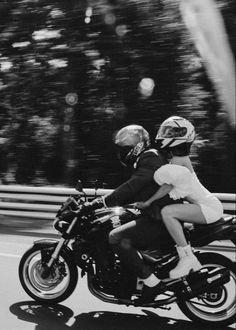 a man and woman riding on the back of a motorcycle down a road with trees in the background