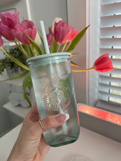 a hand holding a starbucks cup with pink flowers in the window sill behind it