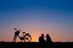 two people sitting next to a bike on top of a grass covered field at sunset