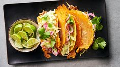 three tacos with limes and cilantro on a black plate