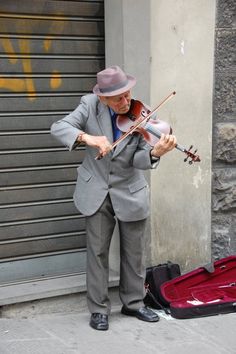 a man in a suit and hat playing the violin on the street next to a suitcase