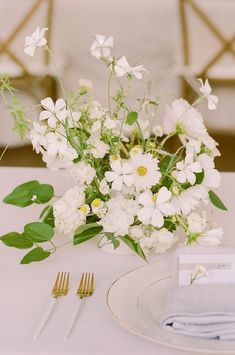 white flowers and greenery in a glass vase on a table with silverware, forks and napkins