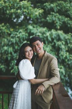 a man and woman standing next to each other in front of some trees smiling at the camera