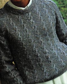 a man wearing a gray sweater and white pants standing in front of a stone wall