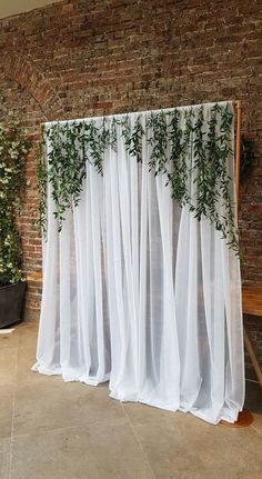 a white curtain with green plants on it in front of a brick wall and wooden bench