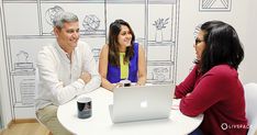 three people sitting at a table talking to each other in front of a laptop computer