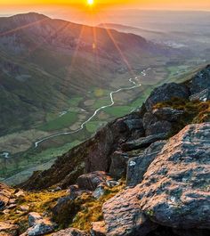 the sun is setting on top of a mountain with rocks and grass in the foreground