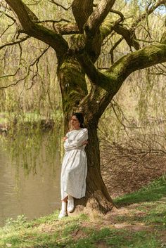 a woman in a white dress leaning against a tree next to a body of water