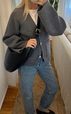 a woman taking a selfie with her cell phone and wearing blue jeans, a gray cardigan sweater and black shoes