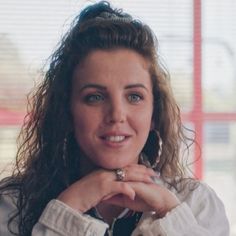 a woman with curly hair wearing a white shirt and silver earrings is looking at the camera