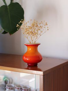 an orange vase with flowers in it sitting on a table next to some cups and a plant