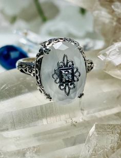 This filigree ring is an Edwardian antique reproduction in sterling silver set with a camphor glass with a sterling silver filigree center embellishment inset with your choice of white CZ, Moissanite, or genuine diamond. The inset center gem is 1mm to 1.5mm in diameter. This faceted oval gem is 14mm x 10mm. This ring sits 7mm off the finger. The inside of the band is marked 925 for solid sterling silver. Ornate Diamond White Filigree Jewelry, Ornate Filigree Diamond White Jewelry, Ornate Diamond White Jewelry With Filigree, White Diamond Crystal Ring With Accent Stones, Diamond White Oval Filigree Jewelry, Silver Filigree Ring With Stone Setting For Formal Occasions, Silver Filigree Ring For Formal Occasion, Silver Crystal Ring With Accent Stones For Jewelry Making, Formal Silver Moonstone Ring With Round Cut