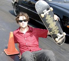 a man sitting on the ground with his skateboard in front of him and a car behind him