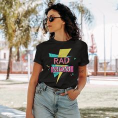 "This totally tubular Rad Mom shirt is perfect for moms who love the 80s or 90s. The retro graphic style and Rad Mom text makes a perfect gift for Mothers Day or mom's birthday! Available on a comfy unisex tee or women's cut tee. We also offer this design on a mug and sweatshirt, and we have a matching Rad Dad design! Check out all our \"rad\" themed items: https://www.etsy.com/shop/LifeLikeDesignsShop?search_query=rad Unisex T-Shirt: Gildan Softstyle .: 100% Cotton (fiber content may vary for d Trendy Mother's Day T-shirt With Graphic Print, Trendy Mother's Day Graphic Print T-shirt, Trendy Mother's Day Graphic T-shirt, Trendy Graphic Print T-shirt For Mother's Day, Retro Black Tops With Funny Text, Retro Black Top With Funny Text, Trendy Graphic Print Top For Mother's Day, Trendy Custom Print Tops For Mother's Day, 80s Mom