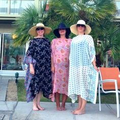 three women standing next to each other in front of a palm tree wearing hats and dresses