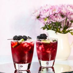 two glasses filled with drinks sitting on top of a glass table next to purple flowers