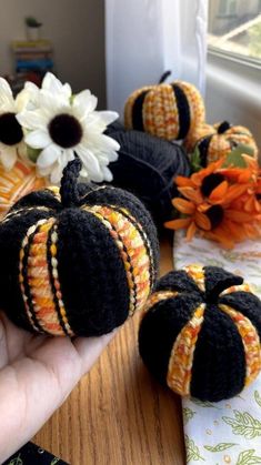 a hand is holding some crocheted pumpkins on a table with flowers in the background