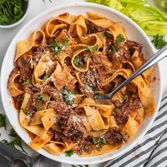 a white bowl filled with pasta and meat on top of a striped cloth next to silverware