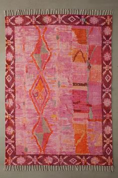 pink and orange rug with fringes hanging on the wall in front of a gray wall