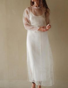 a woman standing in front of a wall wearing a white dress with sheer sleeves and heels