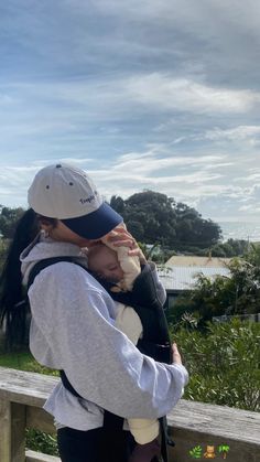 a woman holding a baby in her arms while standing on a wooden bench next to the ocean