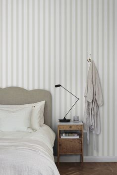 a bedroom with striped wallpaper and white bedding, an end table with a lamp on it