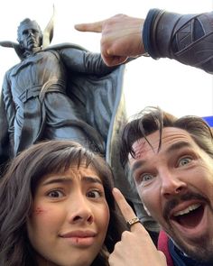 two people standing next to a statue pointing at the camera with their fingers in the air