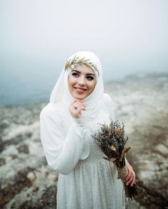 a woman wearing a white hijab and holding flowers in her hand on top of a rocky cliff