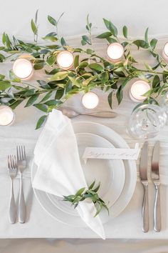 the table is set with white plates and silverware, greenery on each plate