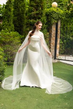 a woman in a white wedding dress and veil standing on the grass with her hands behind her back