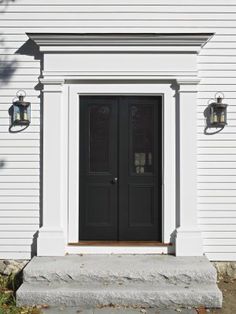 a white house with two black doors and some lights on the side of the building