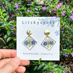 a hand holding up a pair of blue and white flowered earrings in front of purple flowers