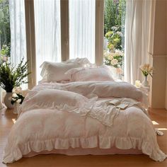 a bed with white ruffled sheets and pillows in front of two large windows that look out onto the garden