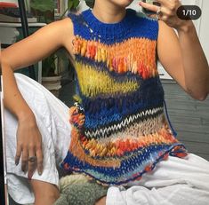 a woman sitting on a bed holding a cell phone up to her face and wearing a multicolored knitted top