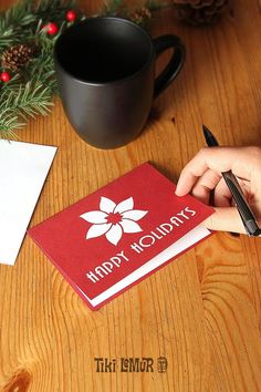 a person writing on a holiday card next to a cup of coffee and pine cones