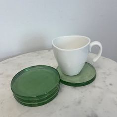 two green glass coasters sitting on top of a white marble table next to a cup and saucer