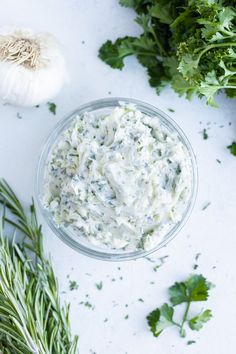 a glass bowl filled with ranch dressing surrounded by fresh herbs and garlic on a white surface