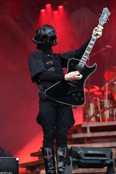 a man with a guitar on stage wearing a black outfit and gas mask holding it up