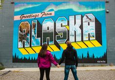 two people holding hands in front of a mural on the side of a building that says alaska