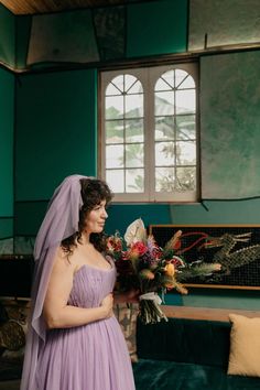 a woman in a purple dress standing next to a green couch and holding a bouquet