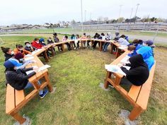 a group of people sitting at wooden benches on top of a grass covered field next to each other